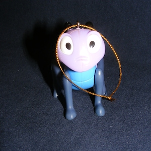 May include: A purple and blue plastic insect figurine with large white eyes and a gold string for hanging. The figurine has a sad expression and is posed on all fours. The background is a dark blue.