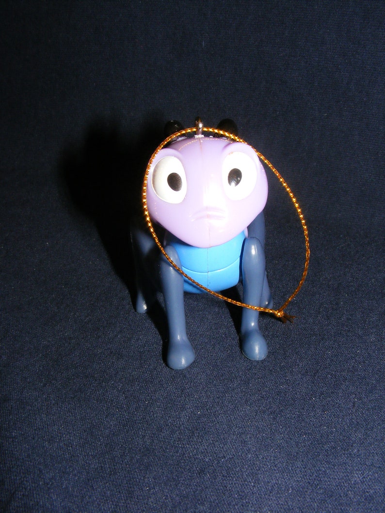 May include: A purple and blue plastic insect figurine with large white eyes and a gold string for hanging. The figurine has a sad expression and is posed on all fours. The background is a dark blue.
