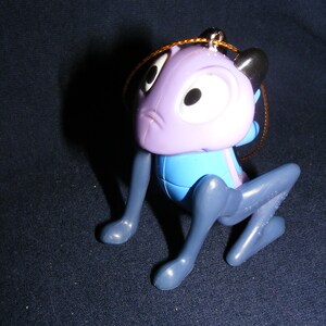May include: A plastic figurine of a cartoon character in a crouching position. The figure has a purple head with large white eyes and black ears. The body is blue, and the legs are gray. A gold string is attached for hanging.