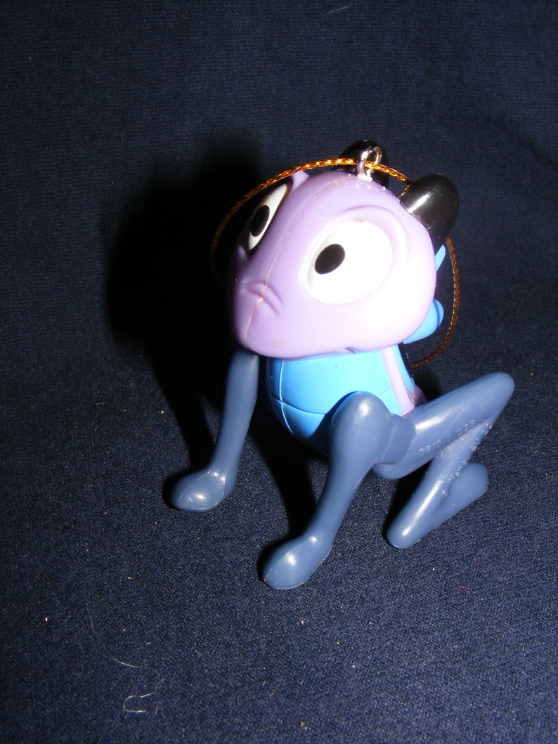 May include: A plastic figurine of a cartoon character in a crouching position. The figure has a purple head with large white eyes and black ears. The body is blue, and the legs are gray. A gold string is attached for hanging.