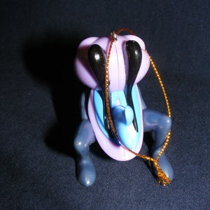 May include: A whimsical ornament featuring a stylized insect-like creature. The ornament has a lavender body, black antennae, and blue accents. It is suspended by a gold-colored string, suggesting it is designed for hanging, possibly on a Christmas tree.
