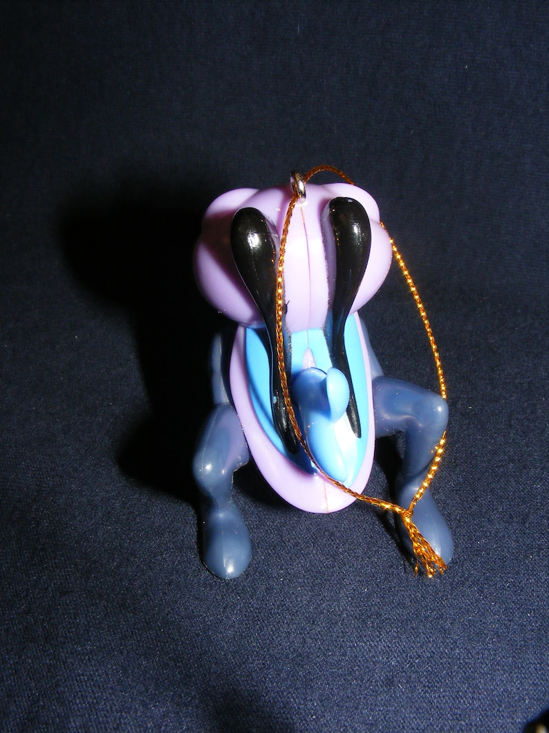 May include: A whimsical ornament featuring a stylized insect-like creature. The ornament has a lavender body, black antennae, and blue accents. It is suspended by a gold-colored string, suggesting it is designed for hanging, possibly on a Christmas tree.