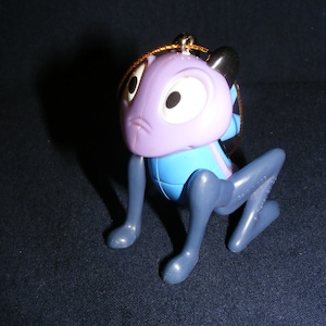 May include: A purple and blue cartoon character ornament with large white eyes and black pupils. The ornament has gray legs and a gold string for hanging. The character is in a crouching position.