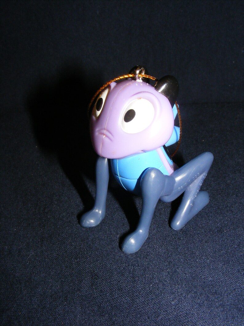 May include: A purple and blue cartoon character ornament with large white eyes and black pupils. The ornament has gray legs and a gold string for hanging. The character is in a crouching position.