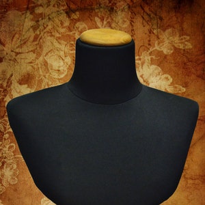 May include: A black fabric mannequin bust with a wooden base. The bust is on a brown background with a floral pattern.