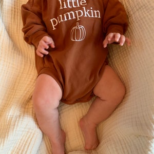 Pu&ograve; includere: Un body per neonato a maniche lunghe marrone con il testo "little pumpkin" e un contorno bianco di una zucca.