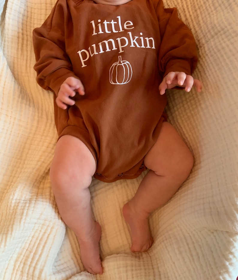 Pu&ograve; includere: Un body per neonato a maniche lunghe marrone con il testo "little pumpkin" e un contorno bianco di una zucca.