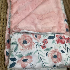 May include: A pink and white floral baby blanket with a soft, plush texture. One side of the blanket is a solid pink color, while the other side features a white background with pink and green floral designs.
