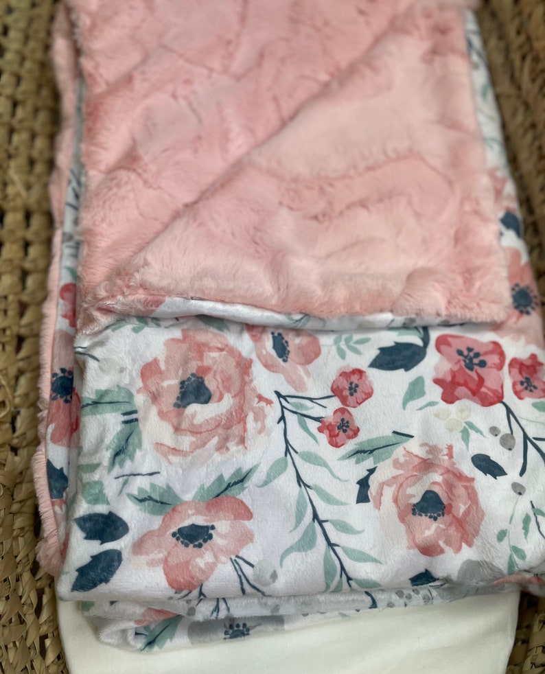 May include: A pink and white floral baby blanket with a soft, plush texture. One side of the blanket is a solid pink color, while the other side features a white background with pink and green floral designs.