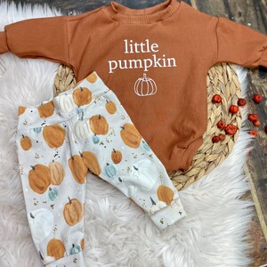 Pu&ograve; includere: Un body per neonato marrone con il testo "little pumpkin" e una piccola grafica di zucca. Il body &egrave; abbinato a pantaloni bianchi con stampa di zucca.