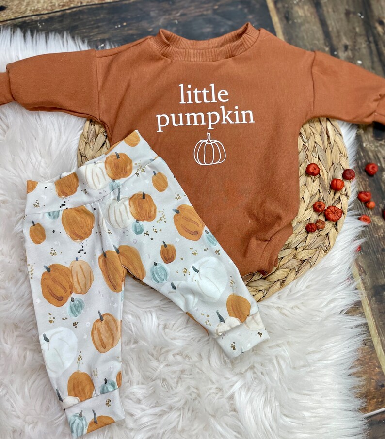 Pu&ograve; includere: Un body per neonato marrone con il testo "little pumpkin" e una piccola grafica di zucca. Il body &egrave; abbinato a pantaloni bianchi con stampa di zucca.