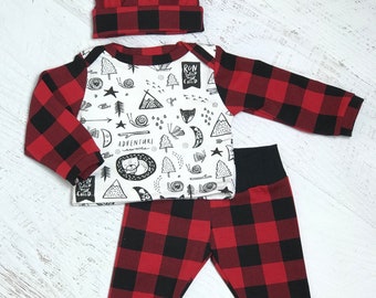 Op de afbeelding: Een driedelige babyoutfitset met een rood en zwart buffelruitpatroon. De set bevat een geknoopte muts, een shirt met lange mouwen met een wit voorpaneel en een bijpassende broek. Het shirt heeft een grafische print met de tekst "Run Wild My Child".