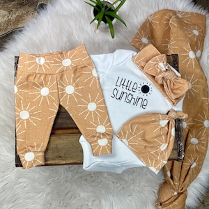 May include: A baby outfit set with a white bodysuit that says "Little Sunshine" with a black sun graphic, a pair of tan pants with white sun graphics, a tan headband with a bow, and a tan hat with white sun graphics.
