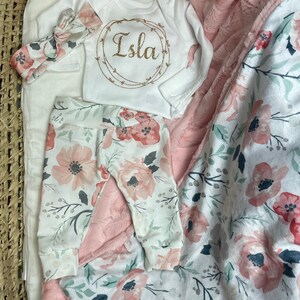 May include: A baby outfit set with a white onesie with the name Isla in gold glitter lettering, pink floral leggings, and a pink floral blanket with a soft pink faux fur lining.