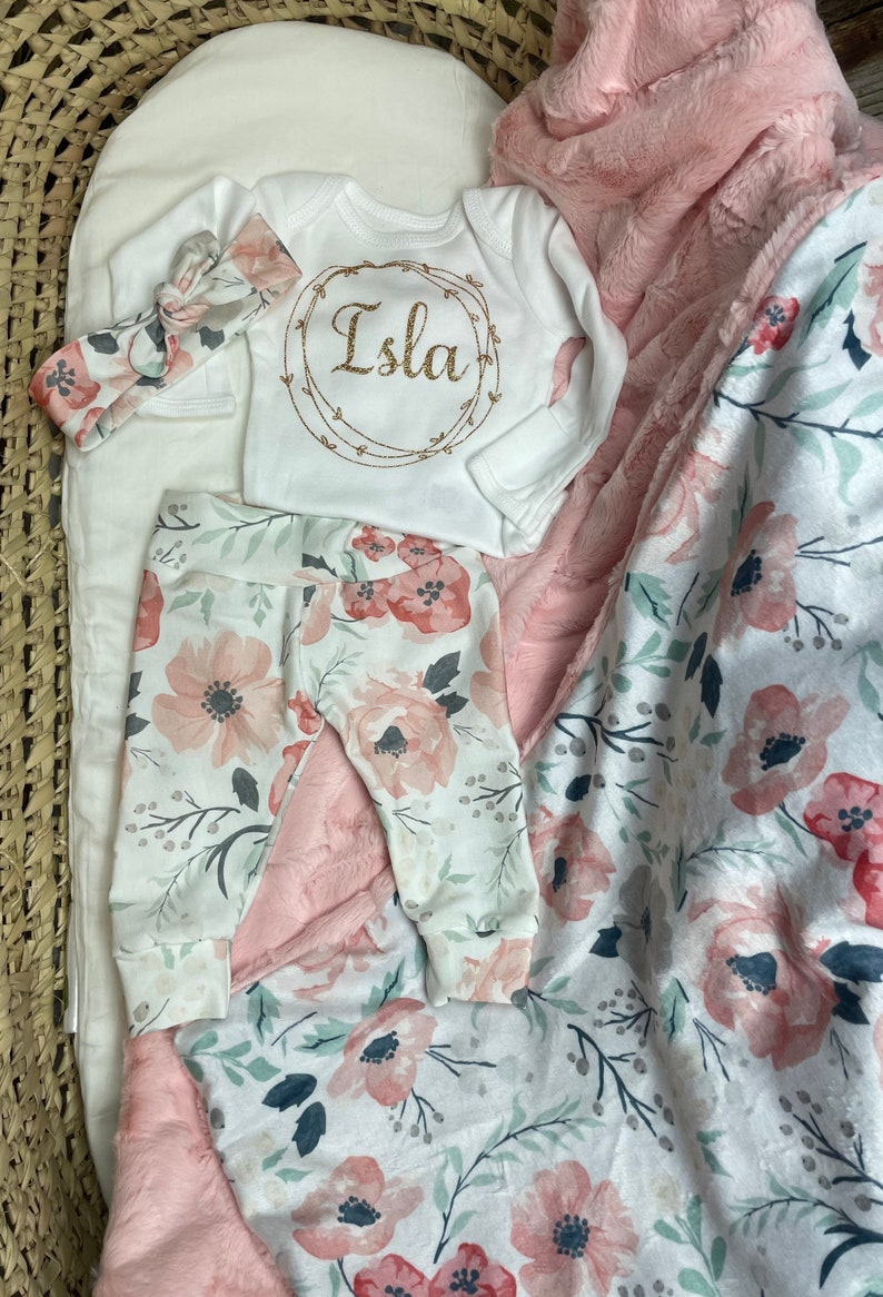 May include: A baby outfit set with a white onesie with the name Isla in gold glitter lettering, pink floral leggings, and a pink floral blanket with a soft pink faux fur lining.