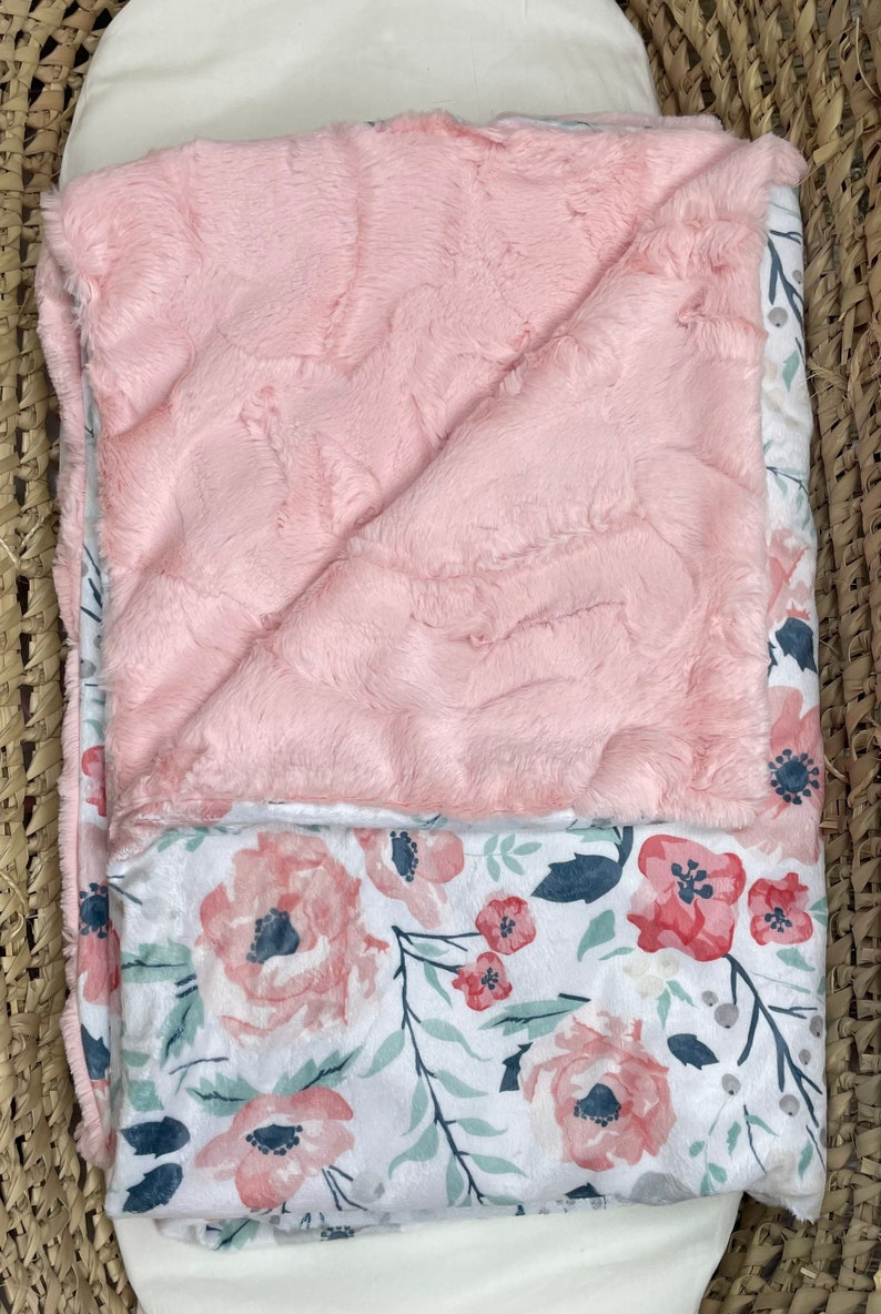 May include: A pink and white baby blanket with a floral pattern. One side of the blanket is soft and fuzzy, while the other side is made of a smooth fabric.