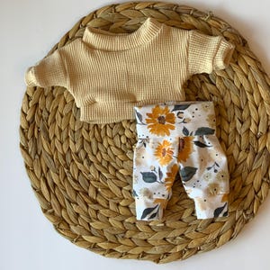 May include: A baby outfit set with a yellow ribbed sweater and white pants with a sunflower print.