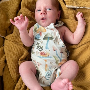 May include: A baby wearing a white onesie with a mushroom and snail print. The onesie has a brown strap and the baby is lying on a yellow blanket.