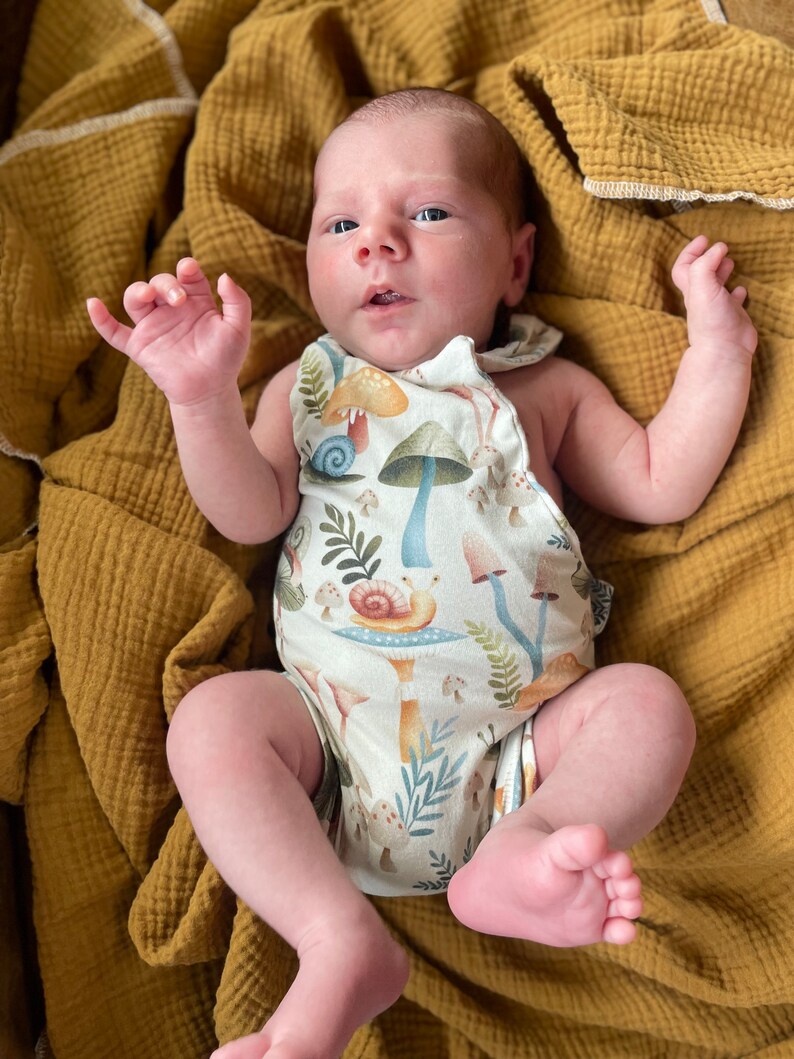 May include: A baby wearing a white onesie with a mushroom and snail print. The onesie has a brown strap and the baby is lying on a yellow blanket.