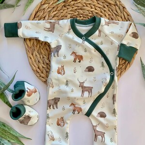 May include: A white baby onesie with a green trim and a woodland animal print. The onesie has a snap closure on the shoulder and a wrap-around design. The print features deer, foxes, owls, squirrels, bears, and hedgehogs.