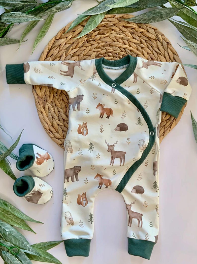 May include: A white baby onesie with a green trim and a woodland animal print. The onesie has a snap closure on the shoulder and a wrap-around design. The print features deer, foxes, owls, squirrels, bears, and hedgehogs.