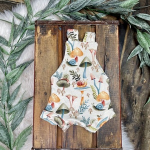 May include: A white baby onesie with a mushroom and snail print. The onesie has a brown strap and is made of soft, comfortable fabric.
