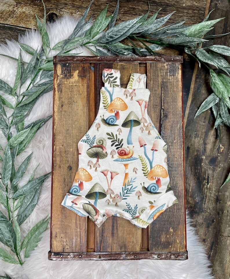 May include: A white baby onesie with a mushroom and snail print. The onesie has a brown strap and is made of soft, comfortable fabric.