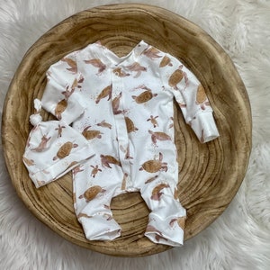 以下が含まれることがあります： 白のベビーロンパースと、お揃いの結び目付き帽子セット。茶色のウミガメと茶色の斑点の繰り返し模様が特徴です。ロンパースは長袖で、前面にスナップが付いています。帽子は上部が結ばれています。