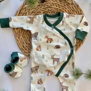 May include: A white baby onesie with a green trim and a woodland animal print. The onesie has a wrap-around design and features bears, deer, owls, squirrels, and hedgehogs. The onesie is paired with a set of green and white booties.