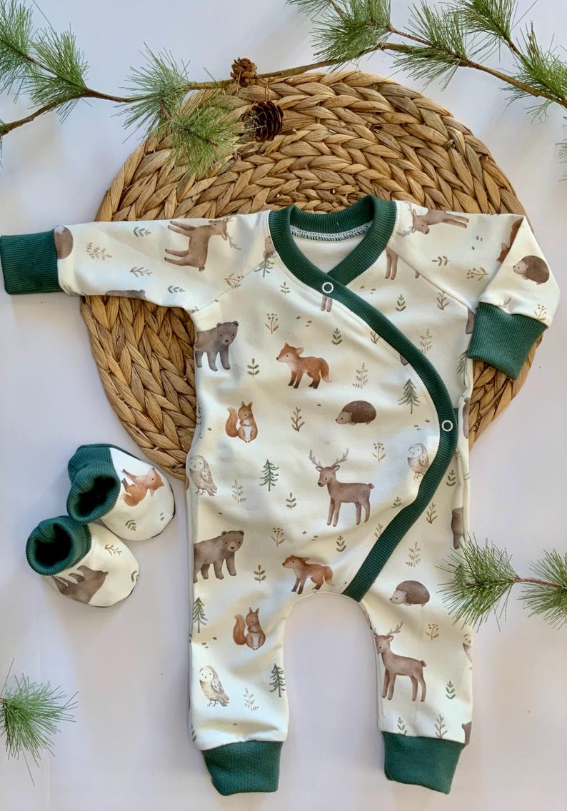 May include: A white baby onesie with a green trim and a woodland animal print. The onesie has a wrap-around design and features bears, deer, owls, squirrels, and hedgehogs. The onesie is paired with a set of green and white booties.