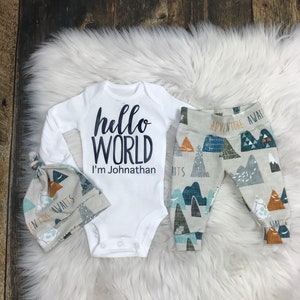 May include: A white baby onesie with the text "hello world I'm Johnathan" in black. A matching hat and trousers with a mountain and adventure theme in shades of blue, brown, and grey.