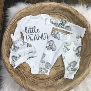 May include: A white baby outfit set with an elephant print. The set includes a long-sleeved onesie with the text "little peanut", a matching hat, and leggings. The headband is also white with an elephant print.