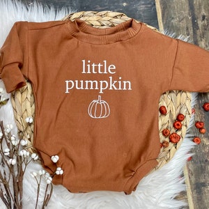Pu&ograve; includere: Un body a maniche lunghe marrone con il testo "little pumpkin" e una piccola grafica di zucca in bianco.