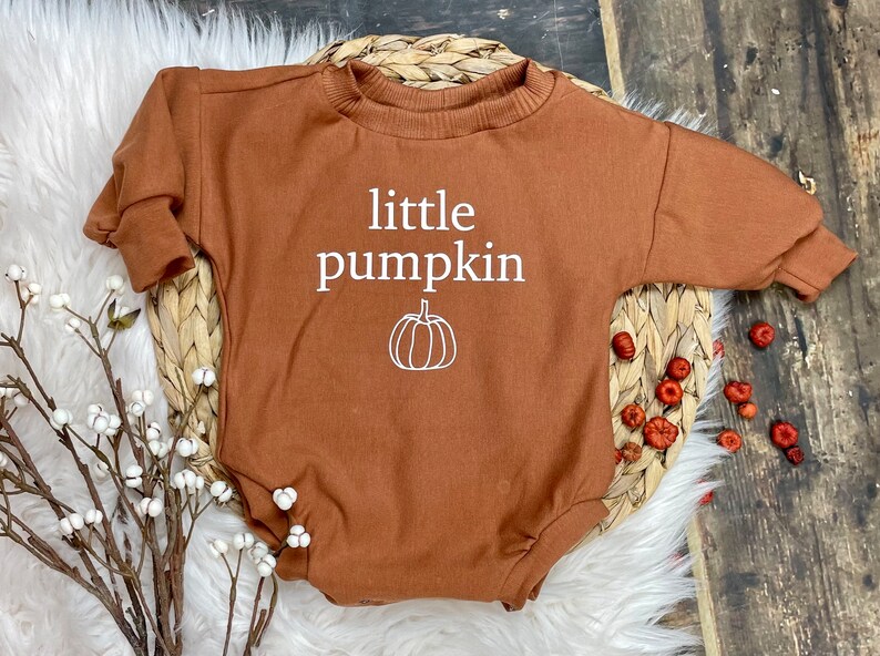 Pu&ograve; includere: Un body a maniche lunghe marrone con il testo "little pumpkin" e una piccola grafica di zucca in bianco.
