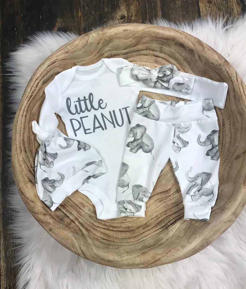 May include: A white baby outfit set with an elephant print. The set includes a long-sleeved onesie, pants, a hat, and a headband. The onesie has the text "little peanut" printed on it.