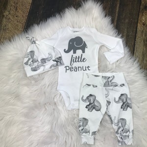K&ouml;nnte beinhalten: Ein wei&szlig;es Baby-Outfit-Set mit Elefanten-Druck. Das Set beinhaltet einen Langarm-Body, eine Hose und eine M&uuml;tze. Der Body hat ein graues Elefanten-Motiv und den Text "little peanut".