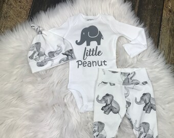 May include: A white baby outfit set with an elephant print. The set includes a long-sleeve onesie, pants, and a hat. The onesie has a gray elephant graphic and the text "little peanut".