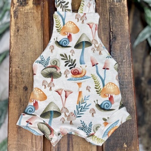 May include: A white baby onesie with a mushroom and snail pattern. The onesie has a light brown background with colorful mushrooms, snails, and leaves.
