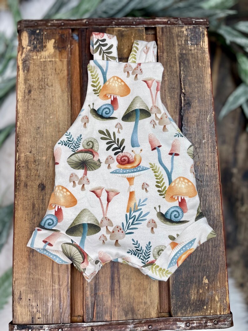May include: A white baby onesie with a mushroom and snail pattern. The onesie has a light brown background with colorful mushrooms, snails, and leaves.