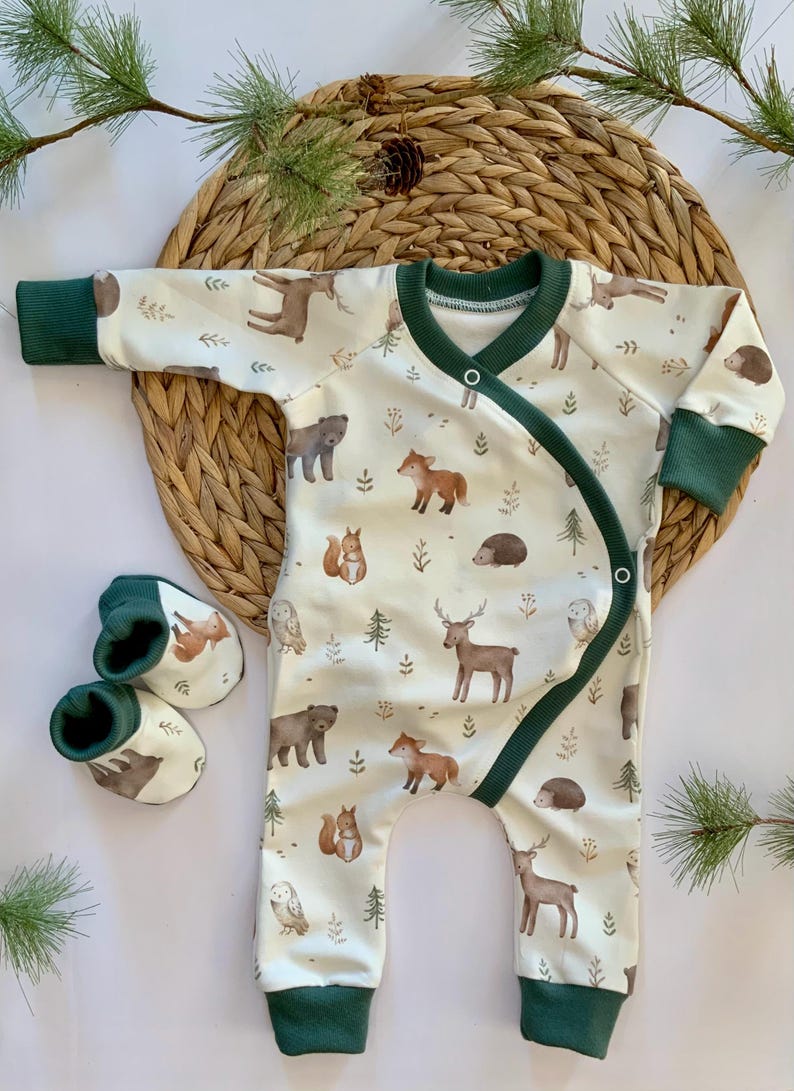 May include: A white baby onesie with a green trim and a woodland animal print. The onesie has a crossover front and is made of soft, comfortable fabric. The onesie is paired with matching green and brown booties.