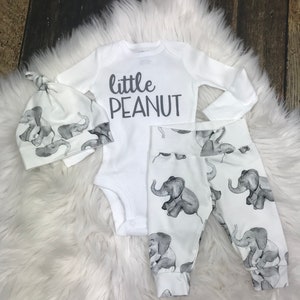 May include: A white baby outfit with an elephant print. The outfit includes a long-sleeved bodysuit with the text "little peanut", a hat, and pants.