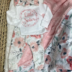 May include: A white baby onesie with the text "Little Sister" in a pink circle, a pink floral headband, and white floral leggings. The outfit is laid out on a pink and white floral blanket.