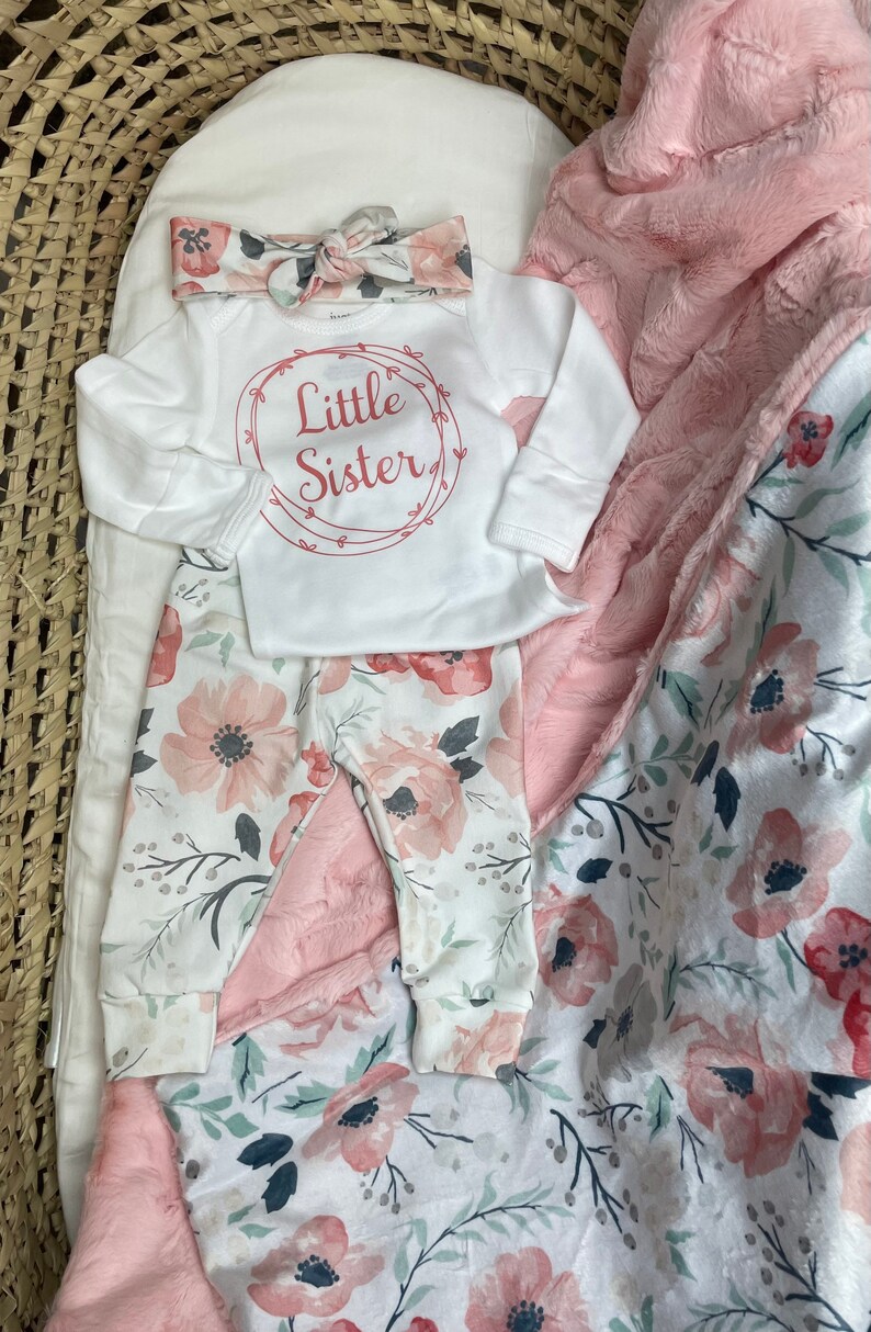 May include: A white baby onesie with the text "Little Sister" in a pink circle, a pink floral headband, and white floral leggings. The outfit is laid out on a pink and white floral blanket.