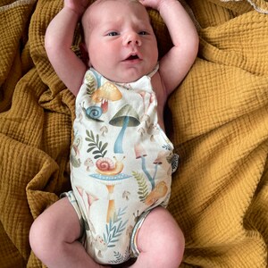 May include: A newborn baby wearing a white onesie with a mushroom and snail print. The onesie has a halter neck and is sleeveless. The baby is lying on a mustard yellow blanket.