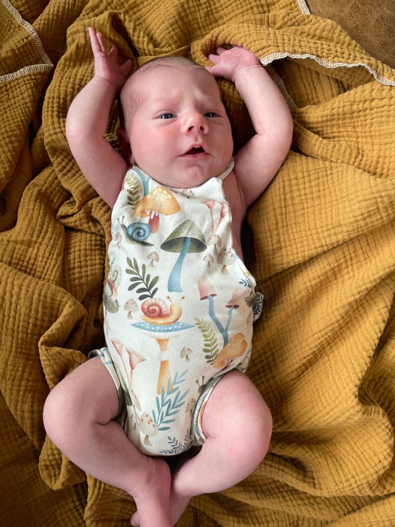 May include: A newborn baby wearing a white onesie with a mushroom and snail print. The onesie has a halter neck and is sleeveless. The baby is lying on a mustard yellow blanket.