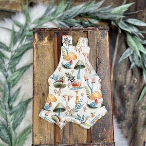 May include: A white baby onesie with a mushroom and snail print. The onesie has a light brown background with colorful mushrooms and snails.