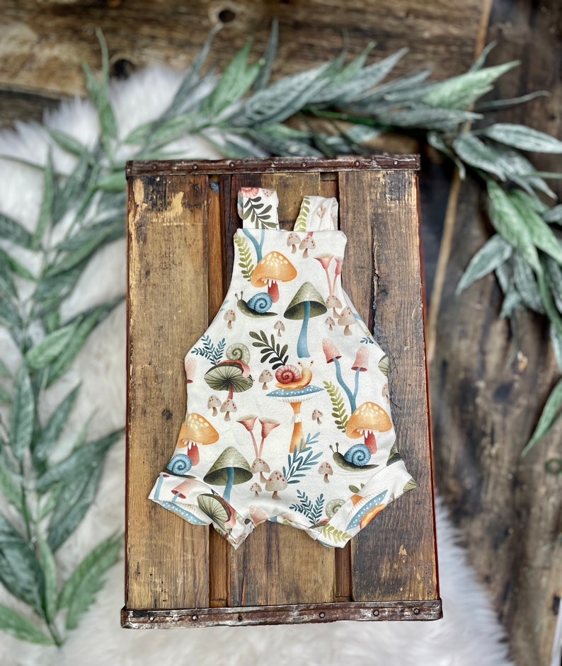 May include: A white baby onesie with a mushroom and snail print. The onesie has a light brown background with colorful mushrooms and snails.