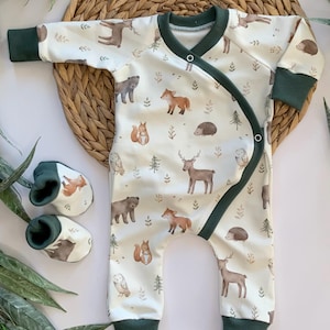 May include: A white baby onesie with a woodland animal print. The onesie has green trim and a wrap-around closure. The print features deer, foxes, owls, squirrels, bears, and hedgehogs.