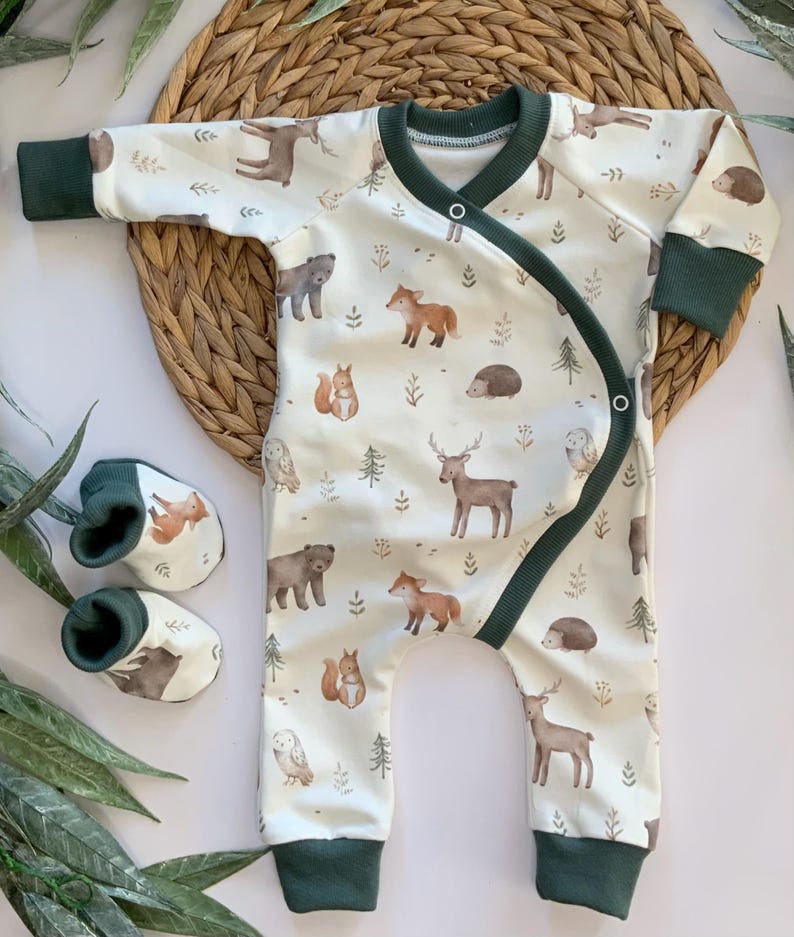 May include: A white baby onesie with a woodland animal print. The onesie has green trim and a wrap-around closure. The print features deer, foxes, owls, squirrels, bears, and hedgehogs.