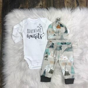 May include: A white baby onesie with the text "A adventure awaits" in black. A matching hat with the text "Adventure awaits" in black. Beige trousers with a mountain and tree pattern in green, blue, and brown.